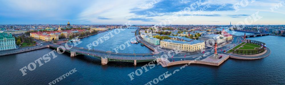 На фотографии изображена панорама Стрелки Васильевского острова с видом на исторические достопримечательности Санкт-Петербурга. Ростральные колонны, украшающие набережную, и величественное здание Биржи занимают центральное место. Река Нева и мосты создают гармоничный пейзаж, а утренний свет подчеркивает архитектурные линии. Этот вид отражает красоту и величие исторического центра города.
